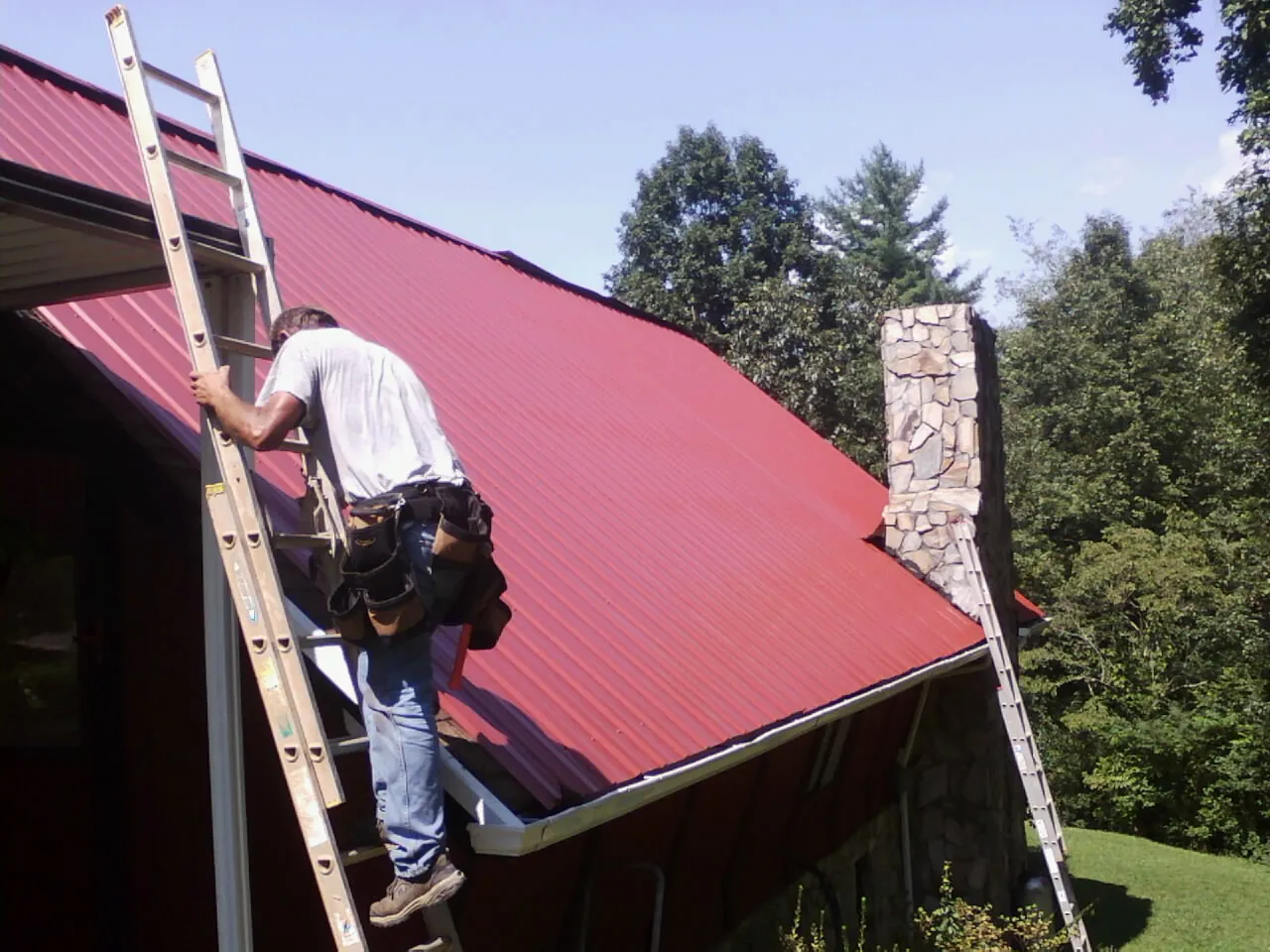 Roofer on a ladder beside a red metal roof and stone chimney — Soffit & Fascia Repair by Roofing Brothers in Noble